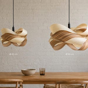 May include: Two wooden pendant lights with a layered, ribbon-like design hang above a wooden table. The larger light is 60 cm in diameter, and the smaller is 42 cm. The lights have a natural wood grain pattern and are suspended from black cords.