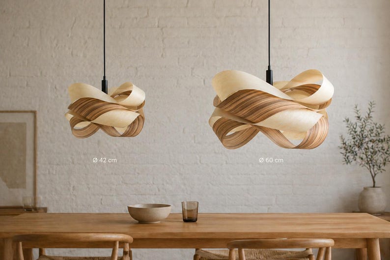 May include: Two wooden pendant lights with a layered, ribbon-like design hang above a wooden table. The larger light is 60 cm in diameter, and the smaller is 42 cm. The lights have a natural wood grain pattern and are suspended from black cords.