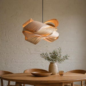 Könnte beinhalten: Eine moderne, geschichtete Holz-Pendelleuchte hängt über einem Holzesstisch. Das Licht hat ein einzigartiges, wirbelndes Design mit warmen, natürlichen Farbtönen. Der Esstisch ist von Holzstühlen und einer Vase mit Grün umgeben.