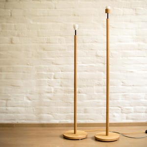 Puede incluir: Dos lámparas de pie de madera con un diseño minimalista. Cada lámpara tiene una base redonda, un poste de madera delgado y una bombilla en la parte superior. Las lámparas están colocadas frente a una pared de ladrillo blanco y un suelo de madera.