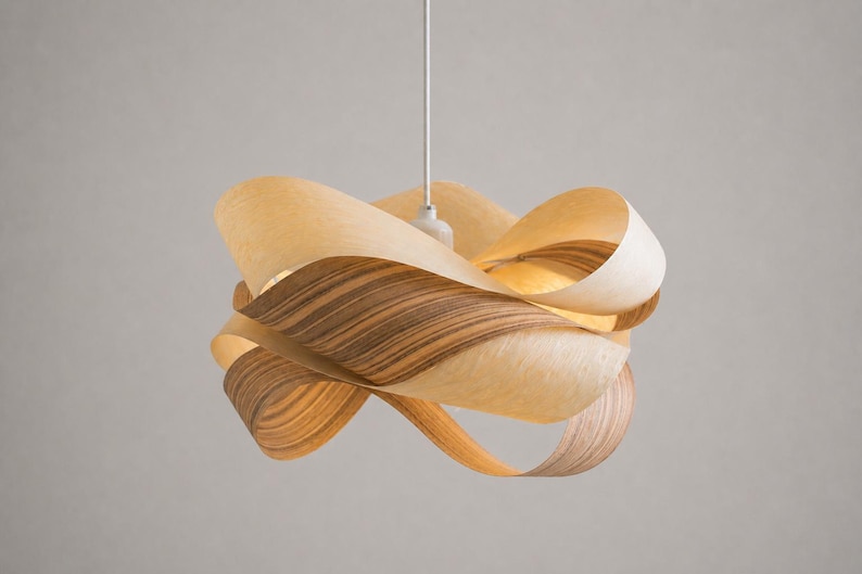 May include: A modern pendant light fixture with a unique, sculptural design. The light is made of layered, curved wood strips in shades of light and dark brown. The fixture hangs from a white cord against a neutral background.