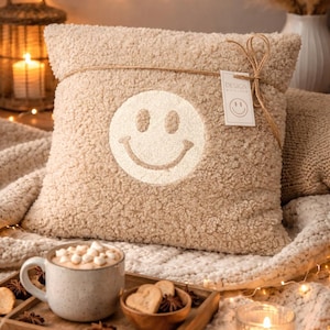 Könnte beinhalten: Beiges, strukturiertes Kissen mit einem großen weißen Smiley-Gesicht. Das Kissen ist mit Bindfaden gebunden und hat ein kleines Etikett. Eine Tasse heiße Schokolade und Kekse stehen auf einem Holztablett vor dem Kissen.