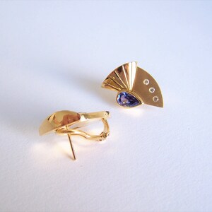 May include: A pair of gold earrings with a purple gemstone and small clear stones. The earrings have a fan-shaped design.