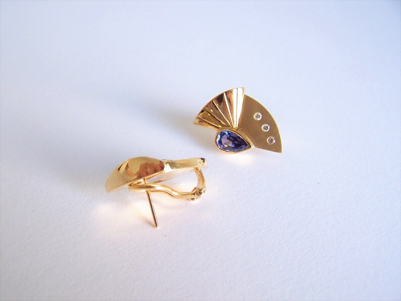 May include: A pair of gold earrings with a purple gemstone and small clear stones. The earrings have a fan-shaped design.