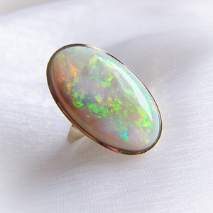 Könnte beinhalten: Ein goldener Ring mit einem großen ovalen Opal-Edelstein. Der Opal hat eine weiße Basis mit grün, blau und pinken irisierenden Blitzen.