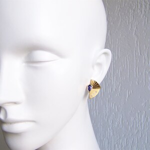 May include: A pair of gold earrings with a purple gemstone and small clear stones. The earrings are shaped like a fan.