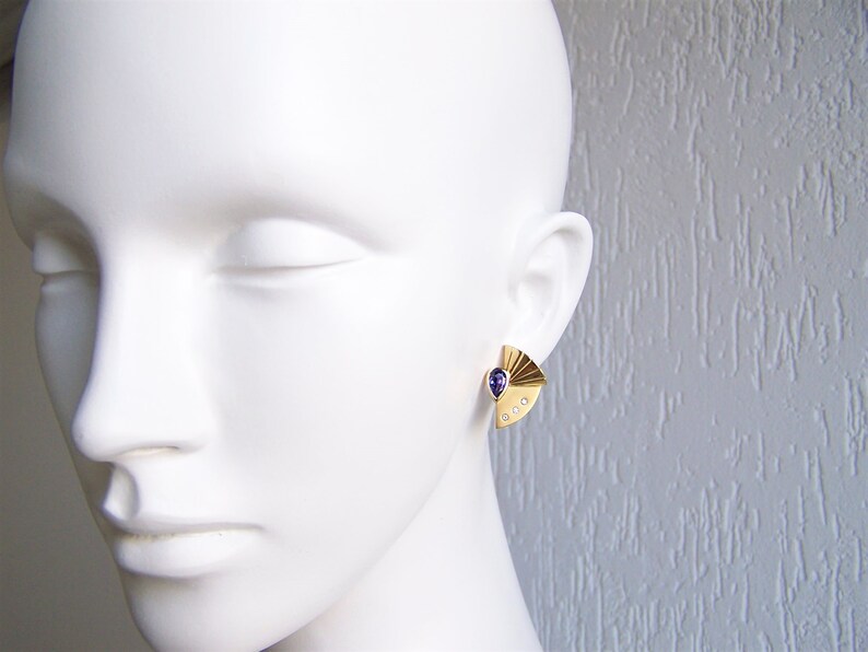 May include: A pair of gold earrings with a purple gemstone and small clear stones. The earrings are shaped like a fan.