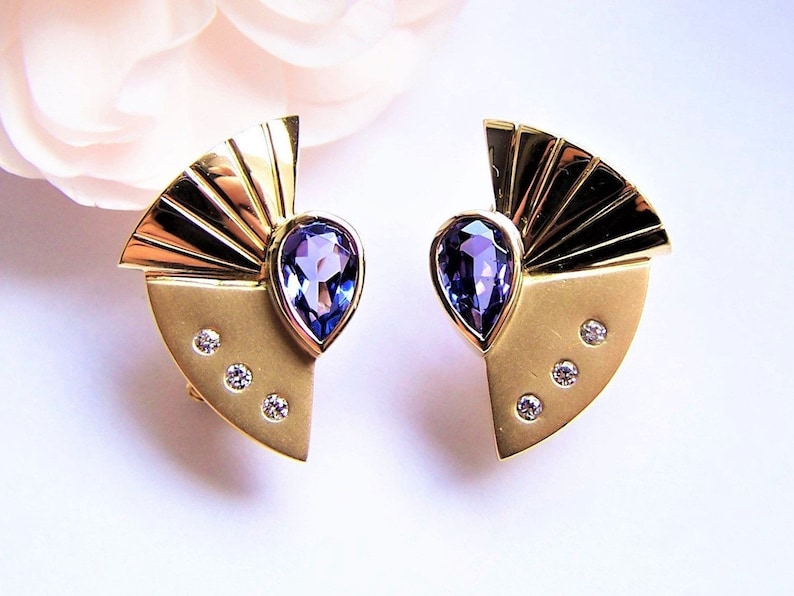 May include: A pair of gold earrings with a fan-shaped design and a purple gemstone in the center. Each earring has three small diamonds on the bottom.
