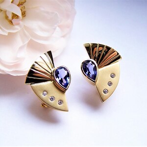 May include: A pair of gold earrings with a fan-shaped design and a teardrop-shaped blue gemstone in the center. Each earring has three small diamonds around the gemstone.