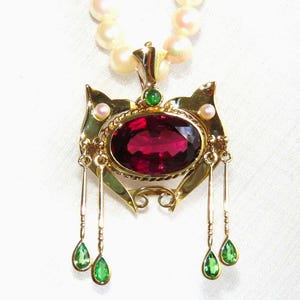May include: A gold-toned pendant necklace featuring a large, oval, deep red gemstone at its center. The pendant is adorned with smaller green and pink gemstones, and teardrop-shaped green gems dangle below. The necklace has a strand of pearls.