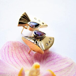 May include: A pair of gold earrings with a geometric design. Each earring features a purple gemstone and small clear gemstones.