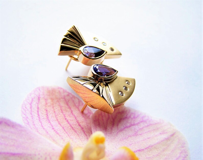 May include: A pair of gold earrings with a geometric design. Each earring features a purple gemstone and small clear gemstones.