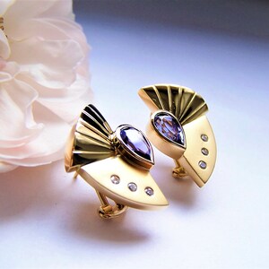 May include: A pair of gold earrings with a fan-shaped design and a pear-shaped amethyst gemstone in the center of each earring. Each earring has three small diamonds around the amethyst.