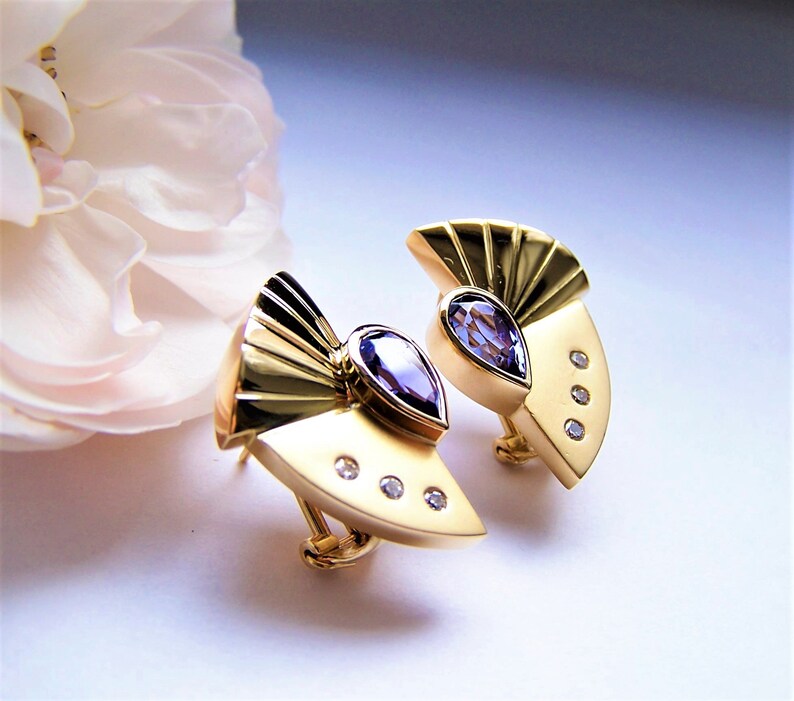May include: A pair of gold earrings with a fan-shaped design and a pear-shaped amethyst gemstone in the center of each earring. Each earring has three small diamonds around the amethyst.