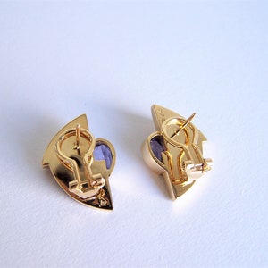 May include: A pair of gold tone earrings with purple stones. The earrings are shaped like a crescent moon and have a hinged closure.