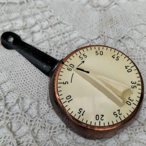 Peut inclure: Un minuteur de cuisine vintage avec une poignée noire et un cadran circulaire. Le cadran a un fond crème avec des chiffres noirs et un bord cuivré. Le minuteur est réglé sur environ 60 minutes.