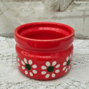 May include: A small, red ceramic container with a cylindrical shape. The container is adorned with a repeating pattern of white daisy-like flowers with green centers. The container sits on a white lace surface.