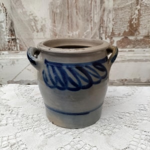 May include: A small, antique stoneware crock with two handles. The gray-blue pot features a blue swirl design around the middle. The interior is dark, and the pot sits on a white lace tablecloth. The background is a distressed, white-painted wooden surface.