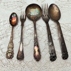 May include: An assortment of antique silverware, including spoons, forks, and a ladle, arranged on a white lace tablecloth. The silverware has ornate designs and a tarnished, vintage appearance, showcasing a classic aesthetic.