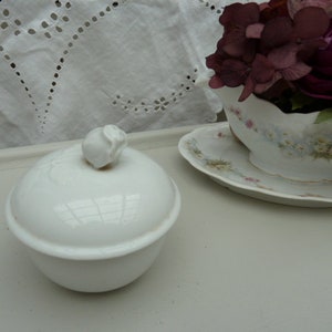 May include: White porcelain sugar bowl with a rosebud lid. The bowl is sitting on a white surface with a white lace tablecloth in the background.