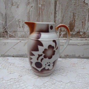 Puede incluir: Una jarra de cerámica vintage con base blanca y diseño floral marrón. La jarra tiene asa y pico marrones. El patrón floral incluye formas de estrella y flor. La jarra está sobre un mantel de encaje blanco.