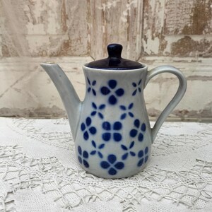 Puede incluir: Una tetera de cerámica con base blanca y estampado floral azul. La tetera tiene tapa y asa azul oscuro. El pico es largo y delgado. La tetera está sobre un mantel de encaje blanco.