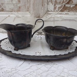 Könnte beinhalten: Ein silbernes Sahnegießer- und Zuckergefäß-Set auf einer Spitzenunterlage. Der Sahnegießer hat einen Griff und einen abgerundeten Körper. Das Zuckergefäß hat einen gewellten Rand und einen runden Körper. Beide Teile sind angelaufen und haben einen Vintage-Look.