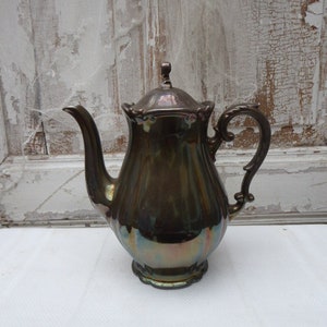 Könnte beinhalten: Eine silberne, irisierende Teekanne mit einem dekorativen Griff und einem Deckel. Die Teekanne steht auf einer weißen Oberfläche.