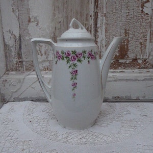 Könnte beinhalten: Weiße Porzellan-Kaffeekanne mit einem Blumendesign aus rosa Rosen und grünen Blättern. Die Kanne hat einen Deckel mit einem Knopf oben.