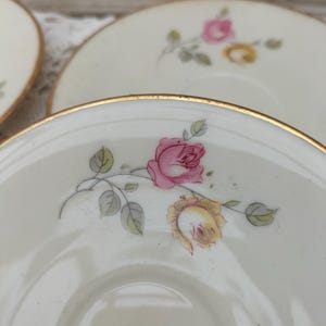 Può includere: Primo piano di diversi piattini biancastri con bordo dorato. Ogni piattino è decorato con un motivo di rose rosa e gialle con foglie verdi. I piattini sono disposti su una superficie di pizzo bianco.