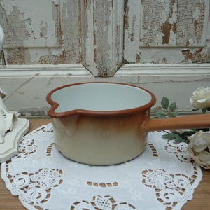 May include: A white enamel saucepan with a brown rim and handle. The saucepan is sitting on a white doily.