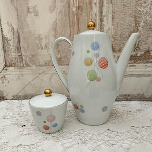 Può includere: Servizio da caffè in ceramica bianca composto da caffettiera e zuccheriera. La caffettiera ha un pomello dorato sul coperchio e un manico curvo. Entrambi i pezzi sono decorati con pois colorati e un design delicato.