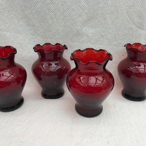 Könnte beinhalten: Vier kleine, tiefrote Glasvasen in einer Reihe angeordnet. Jede Vase hat einen bauchigen Körper, einen schmalen Hals und einen breiten, gerüschten, gewellten Rand. Das Glas ist durchscheinend und glatt und reflektiert das Licht.