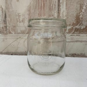 May include: A clear glass jar with a wide mouth and a screw-on lid. The jar has a rounded body and a logo embossed on the front. The jar is sitting on a white surface, with a distressed wooden background.