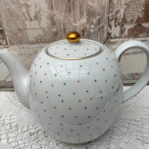 Puede incluir: Tetera de cerámica blanca con cuerpo redondo, asa curva y boquilla. La tapa tiene un pomo y un borde dorados. La tetera está decorada con pequeños patrones de estrellas rojas y negras. Un complemento encantador.