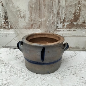 Könnte beinhalten: Kleiner, antiker Steinguttopf mit zwei Henkeln. Der Topf ist hellblau-grau mit einem dunkelblauen Streifen und vertikalem Akzent. Er steht auf einer weißen Spitzentischdecke vor einem verwitterten weißen Holzhintergrund.