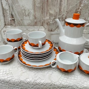 Könnte beinhalten: Vintage weißes Keramik-Teeservice mit orangefarbenen und braunen Muschelmustern. Das Set umfasst eine Teekanne, einen Sahnekännchen, eine Zuckerdose, Tassen und Untertassen. Die Tassen und Untertassen sind gestapelt.