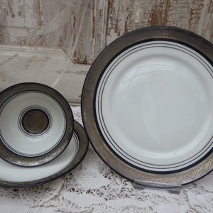 May include: A set of three white ceramic plates with black and gold trim. The plates are arranged in a pyramid shape, with the largest plate at the bottom and the smallest plate at the top.