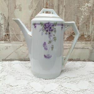 Puede incluir: Cafetera de porcelana blanca con asa y boquilla geométricas. La cafetera está decorada con diseños florales morados y hojas verdes. Un ribete dorado acentúa el borde y el asa. La cafetera está sobre un mantelito de encaje blanco.