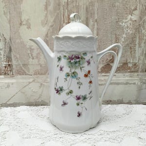 May include: White porcelain coffee pot with a floral design featuring pink, blue, and orange flowers. The pot has a scalloped rim and a lid with a knob.