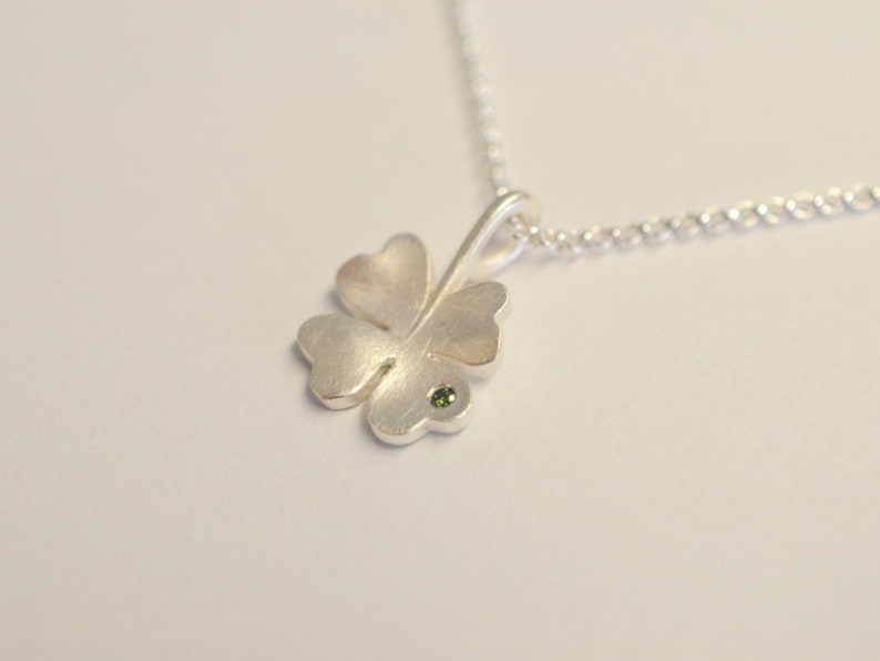 Op de afbeelding: Een zilveren ketting met een vierbladig klavertjevier-hanger, met een klein groen edelsteentje in het midden.