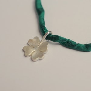 May include: A silver four-leaf clover pendant hangs from a dark green satin ribbon. The clover has a brushed metal finish. The ribbon is twisted and appears to be knotted. The background is a plain white.