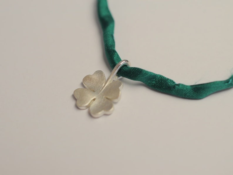 May include: A silver four-leaf clover pendant hangs from a dark green satin ribbon. The clover has a brushed metal finish. The ribbon is twisted and appears to be knotted. The background is a plain white.