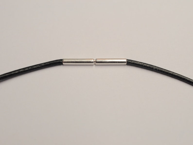 May include: A close-up of a black leather necklace cord with a silver-colored clasp. The cord is round and appears to be approximately 2 mm in diameter. The clasp is made of two cylindrical pieces that connect in the center.