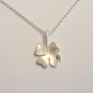 Op de afbeelding: Een zilveren ketting met een klavertje vier hanger met een kleine groene edelsteen.