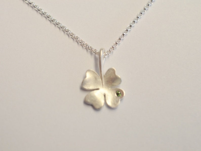 Op de afbeelding: Een zilveren ketting met een klavertje vier hanger met een kleine groene edelsteen.
