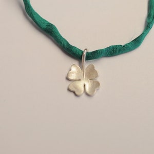 May include: A silver four-leaf clover pendant hangs from a green fabric necklace. The pendant is a detailed, textured design. The necklace is a vibrant green color, and the background is a soft white.