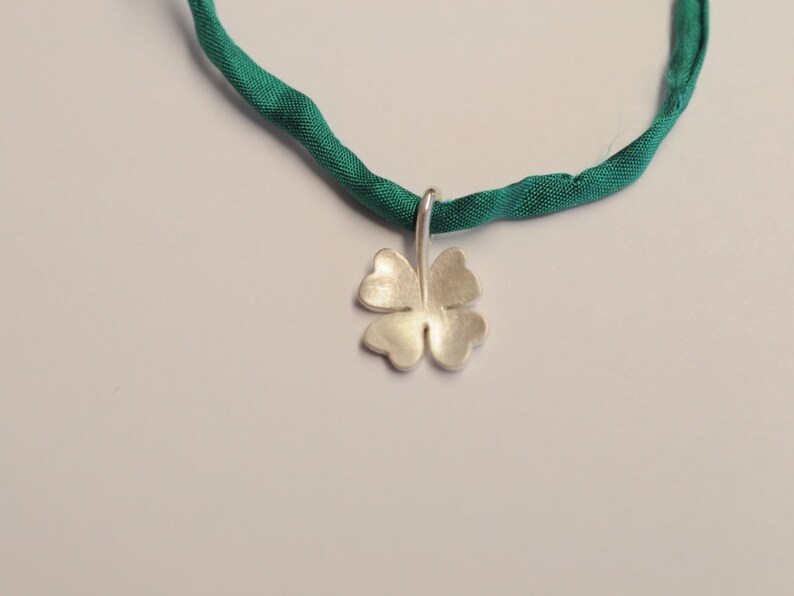 May include: A silver four-leaf clover pendant hangs from a green fabric necklace. The pendant is a detailed, textured design. The necklace is a vibrant green color, and the background is a soft white.
