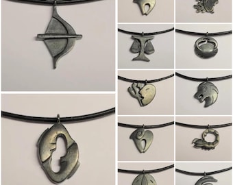 Op de afbeelding: Een collectie van twaalf donkere metalen dierenriemhangers aan zwarte koordkettingen. Elke hanger toont een uniek astrologisch symbool, waaronder een pijl en boog, een weegschaal, een leeuw, een schorpioen en andere gestileerde tekens.
