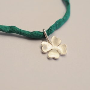 May include: A silver four-leaf clover pendant hangs from a dark green fabric necklace. The clover has a matte finish and is centered in the frame. The necklace is a twisted, textured fabric. The background is a light gray.
